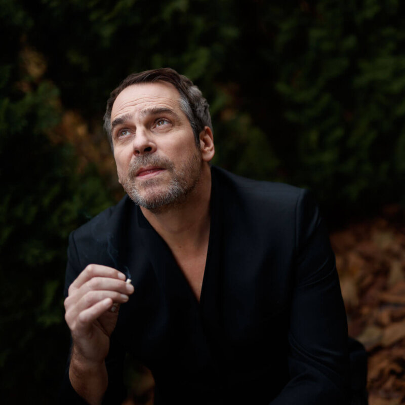 Zdjęcie przedstawia aktora Pawła Deląga w eleganckim stroju, patrzącego lekko w bok. Artystyczne portretowe ujęcie z miękkim światłem i neutralnym tłem.