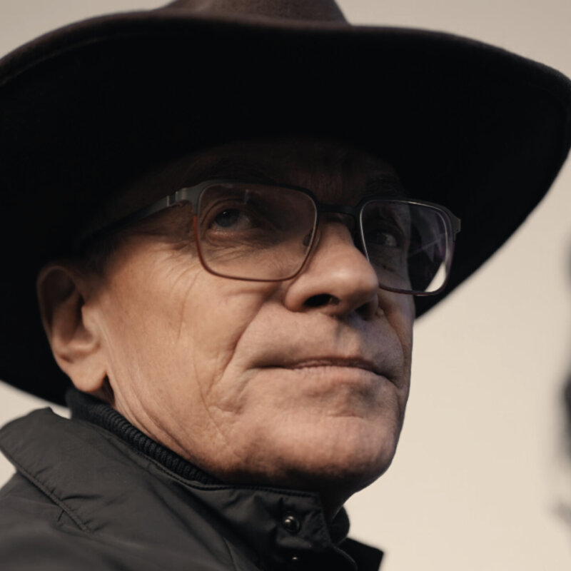 Zbliżenie na głowę mężczyzny w kowbojskiej czapce. Twarz wypełnia kadr, a tło stanowi rozmyta przestrzeń plenerowa.