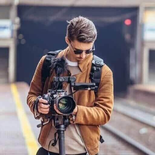 Dariusz Goldmann trzyma kamerę na statywie na stacji kolejowej, patrzy w dół, ubrany w brązową kurtkę i okulary